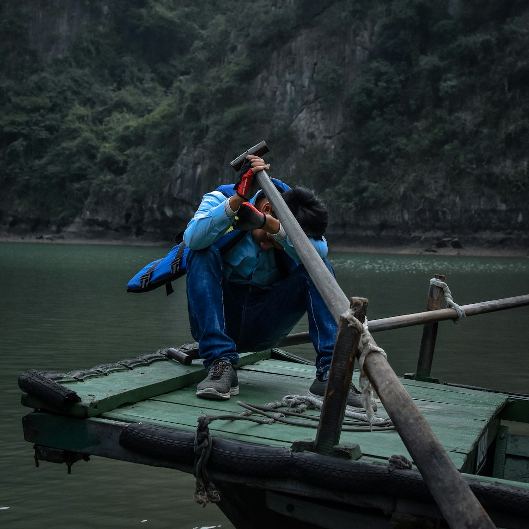man in blue jacket and blue denim jeans sitting on brown wooden dock during daytime